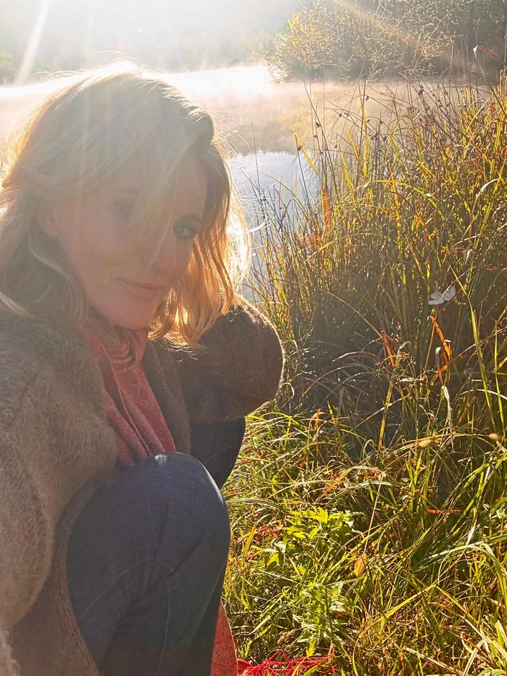 Eine Frau am Ufer eines Sees im warmen Gegenlicht, neben Gräsern und Schilf